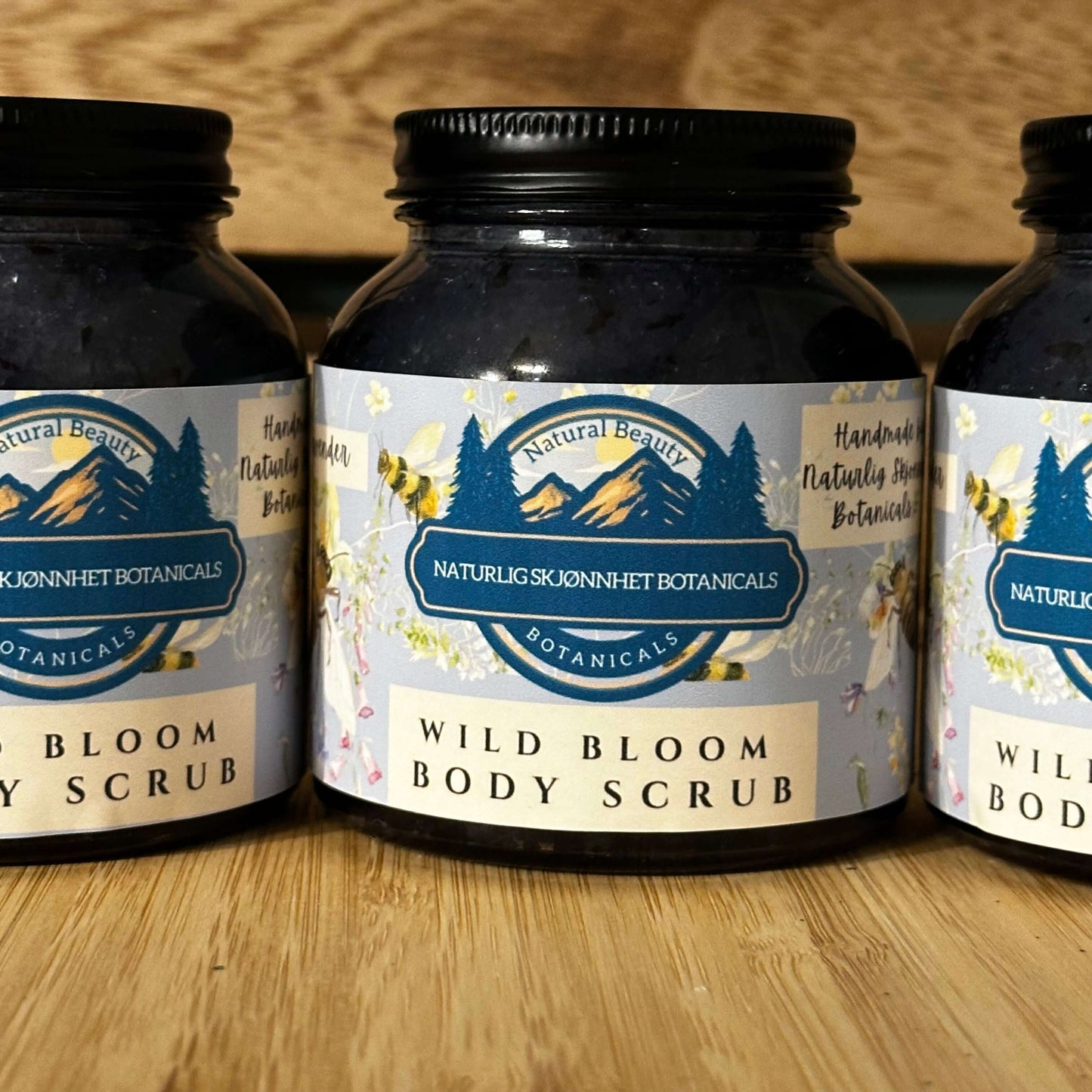 Three jars of Wild Bloom Body Scrub with floral labels, showcasing natural, handcrafted ingredients for smooth skin.